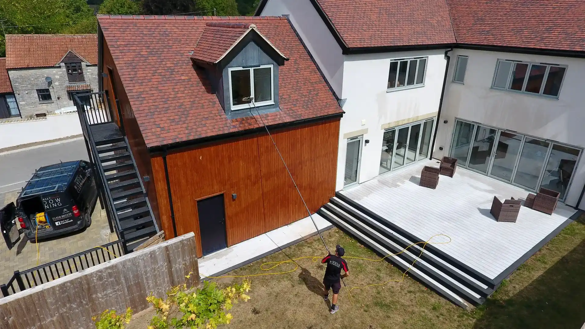 Aerial view of a Somerset Window Cleaning technician cleaning timber-clad and rendered elevations with a reach-and-wash pole, van parked alongside
